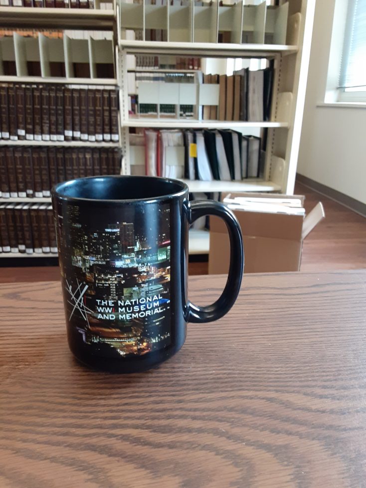 A cup of coffee in a World War One Museum mug is sitting on a table. In the background are some empty library shelves.